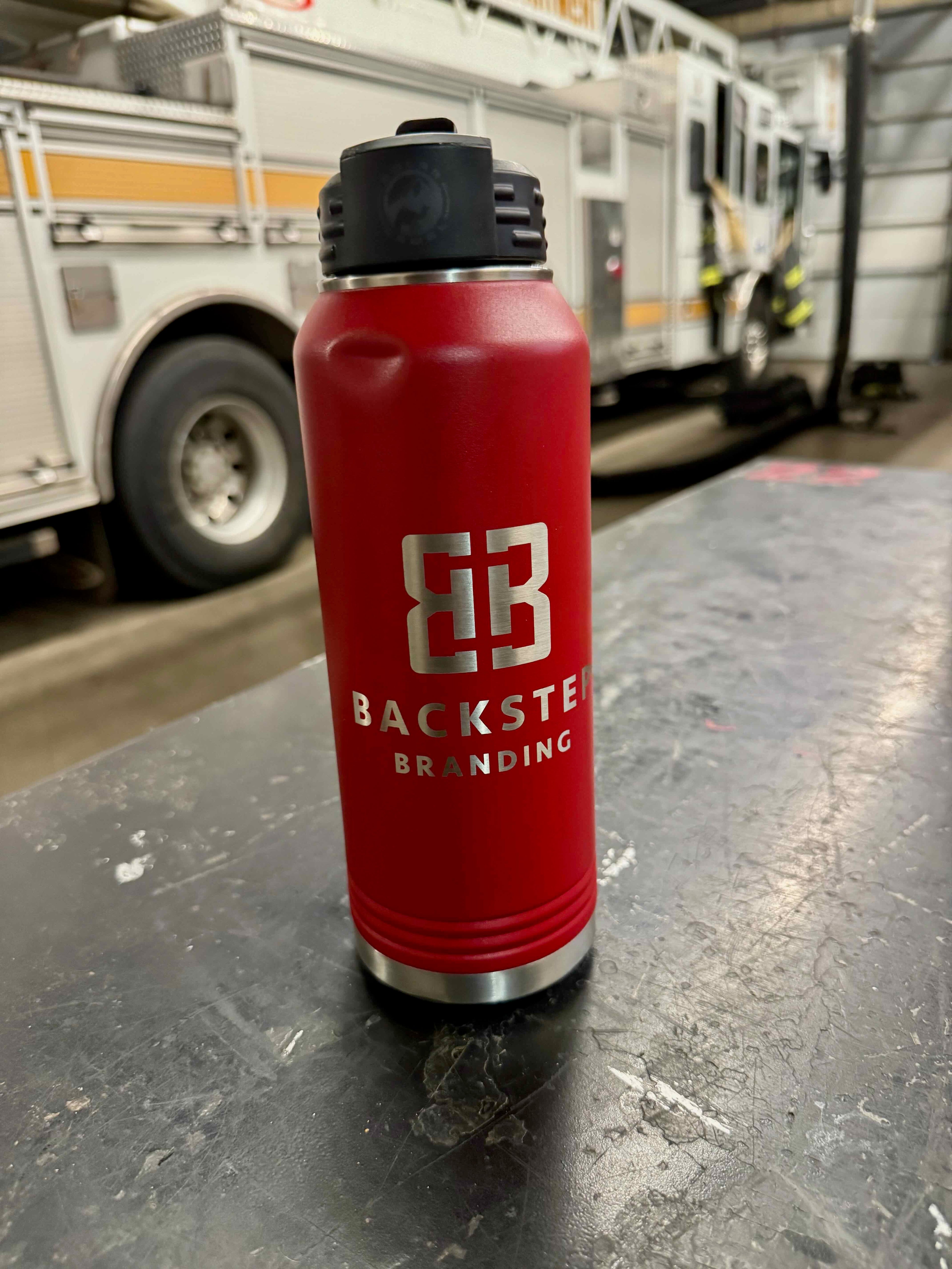 Red water bottle with 'Backsteed Branding' logo on a metallic surface with a fire truck in the background.