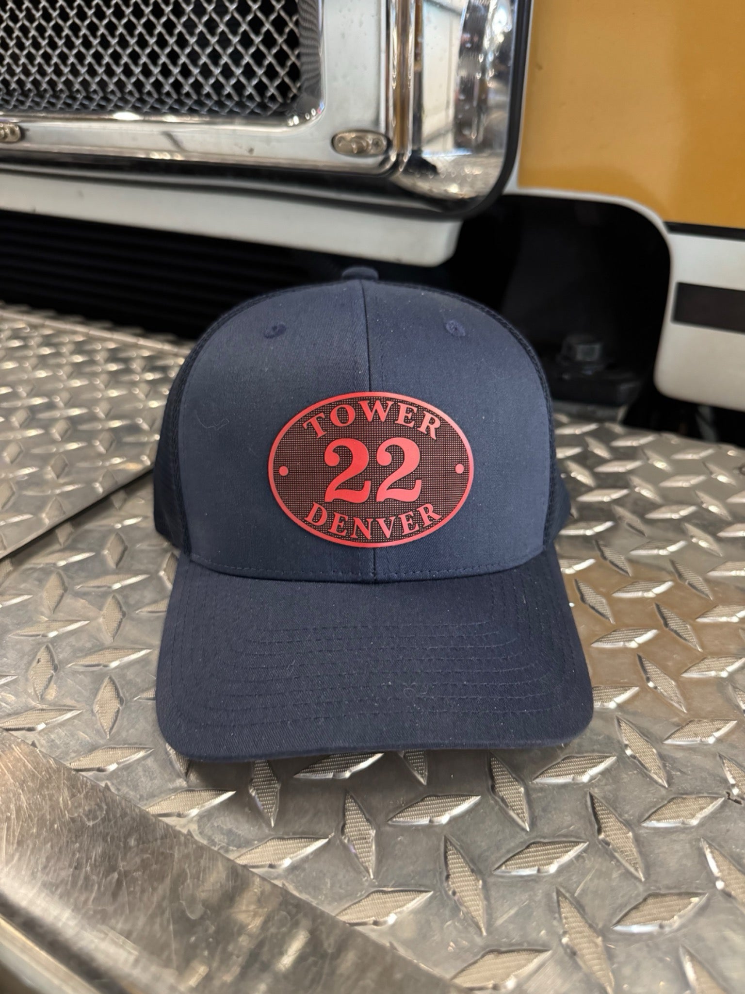 Navy blue cap with red 'Tower 22 Denver' logo on a metallic surface with a vehicle in the background